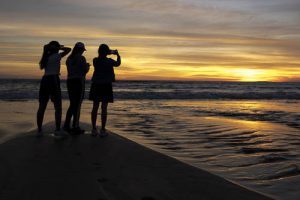 Three people in silhouette as they take photos of a beach sunset